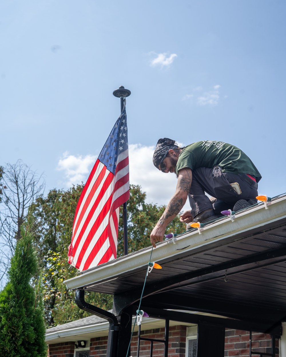 Anderson Lawn Care crew member on a roof clipping orange and purple Halloween bulbs to the gutter next to an American flag