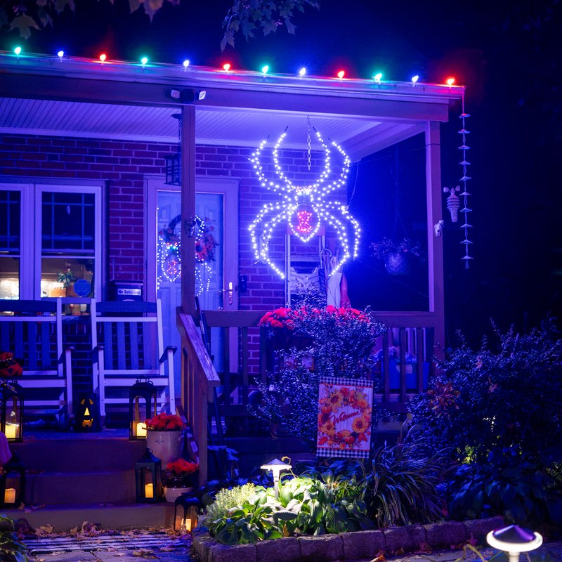 Brick front porch at night with multicolor bulbs along the roofline and a large lit spider for Halloween